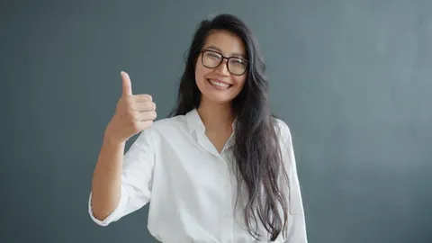 Smiling woman in glasses and white shirt giving a thumbs up against gray background.